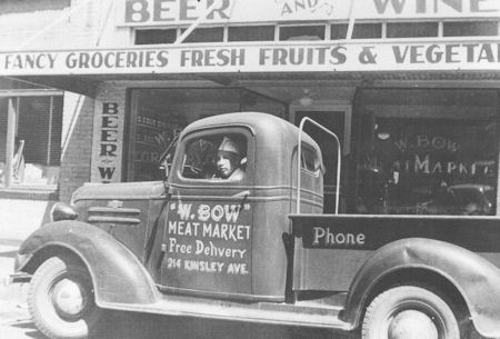 Black-and-white photo of a vintage pickup truck labeled "W. Bow Meat Market" parked in front of a grocery store selling beer, wine, fresh fruits, and vegetables.