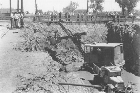 Black and white photo of a construction site with an excavator digging a large pit, a truck nearby, and a group of people watching from the edge.