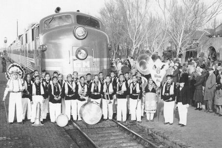 A marching band poses in uniform in front of a stationary train, with a crowd of people gathered on the platform in the background.
