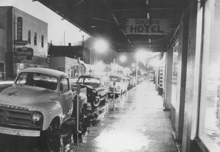 Black and white photo of a wet city street at night with parked cars, illuminated shop windows, and a hotel sign overhead.