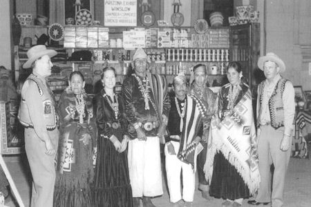 A group of seven people, some in traditional Native American clothing and others in Western clothing, pose in front of a display of crafts and goods.