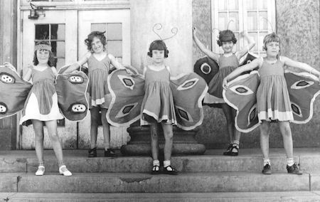 Five young girls wearing butterfly costumes stand on outdoor steps with arms raised, posing for a group photo in front of a building.