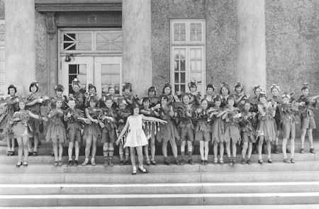 A group of children in costume poses in rows on the steps of a building, with one child in a white dress standing at the front center.