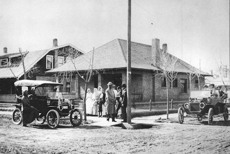 Black and white photo of early 20th-century cars on a dirt road with a group of people standing in front of a house.