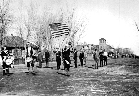 A group of people, some holding drums and an American flag, stand in formation on a dirt road in a small town with houses and trees in the background.