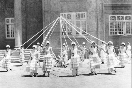 A group of women in tiered dresses and bonnets dance around a maypole, holding ribbons, outside a large building with tall windows and steps.