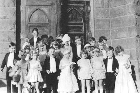 A group of children dressed in formal attire poses in front of large wooden doors and a stone building.