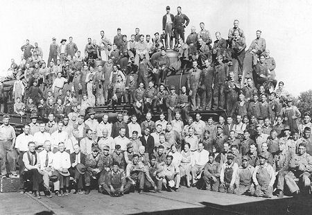 A large group of men pose on and in front of a locomotive engine, wearing work clothes, in a black-and-white historical photo.