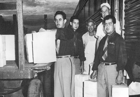 Five men stand beside a train, holding and loading boxes onto a platform. One man wears an apron and hat, while the others are in shirts and slacks.