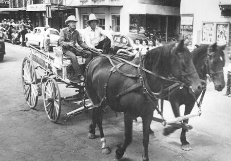 Two men wearing hats ride a horse-drawn wagon down a city street, with people, cars, and storefronts visible in the background.