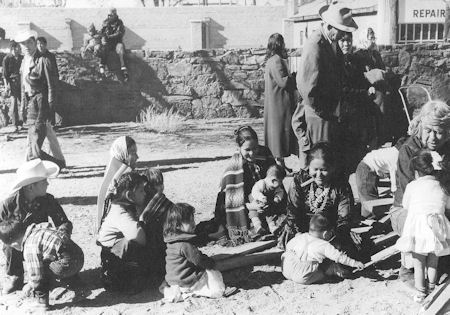 A group of adults and children sit and stand on sandy ground near a stone wall, with some people wearing hats and blankets. A building with a "REPAIR" sign is visible in the background.