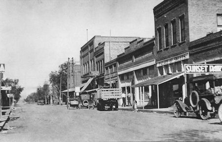 Black and white photo of an early 20th-century street with cars parked along storefronts, including one labeled "Sunset Dairy," and trees lining the road on the left.