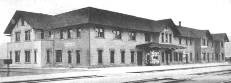 Black-and-white photo of a two-story brick train station building with a covered entrance and visible railroad tracks in the foreground.