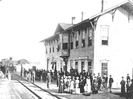 Black and white photo of a train station with a group of people standing on the platform near the tracks, next to a two-story wooden building.