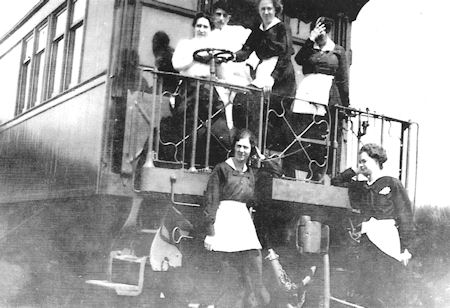 A group of women in uniforms and aprons pose on the steps and platform of a railway carriage in a black and white photo.