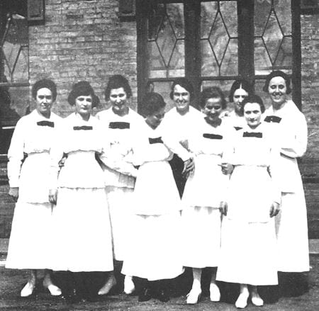 A group of eight women dressed in similar white uniforms with dark bows stand together outside a brick building with large windows.