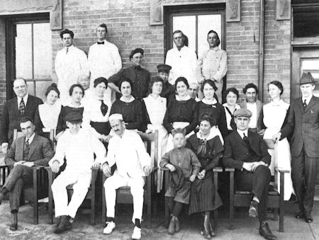 A black-and-white photo of a large group of men, women, and children posing together in front of a brick building. Some are seated, others are standing, and most wear early 20th-century clothing.