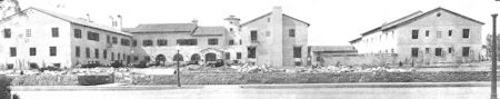Black and white photo of a large building complex with debris scattered in front, surrounded by a low stone wall and a street in the foreground.
