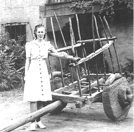 A woman in a light-colored dress stands beside a large wooden cart with spoked sides and solid wheels in an outdoor setting.