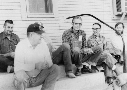 Five men sit on concrete steps outside a wooden building, engaged in conversation and appearing relaxed.