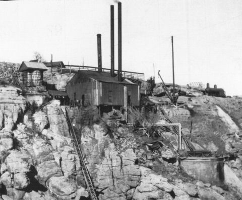 Black and white photo of an old industrial building with two tall smokestacks, perched on a rocky cliff with machinery and equipment nearby.