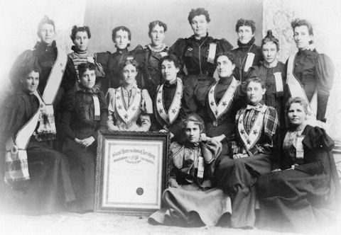 Black and white photo of fifteen women in formal clothing, some seated and some standing, posing with a large framed certificate.