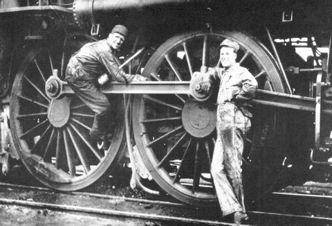 Two men in work clothes pose near the large wheels and rods of a steam locomotive on a railway track.
