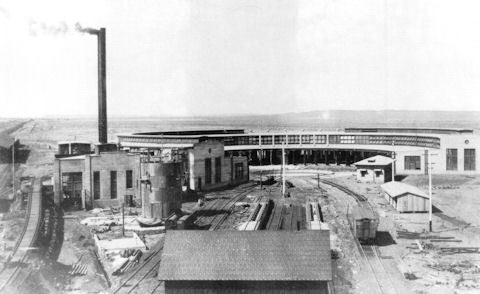 Black and white photo of an early 20th-century industrial railway roundhouse with a smoke stack, tracks, and several utility buildings.