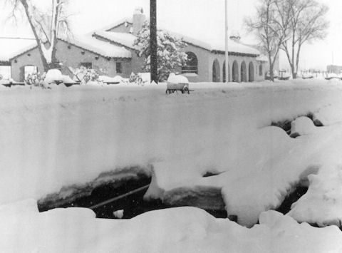 A building stands in the background while deep snow covers the ground, partially burying railroad tracks in the foreground.