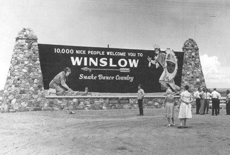 Black-and-white photo of people standing in front of a large stone sign reading, "10,000 nice people welcome you to Winslow, Snake Dance Country.