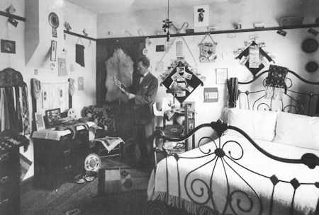 A man in a suit stands in a decorated bedroom, reading a paper. The room has a metal bed, various wall decorations, and a cluttered dresser.