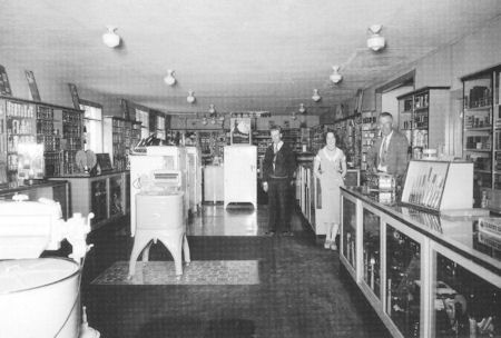 Black and white photo of four people standing in an old-fashioned store with shelves of goods and large appliances on display.