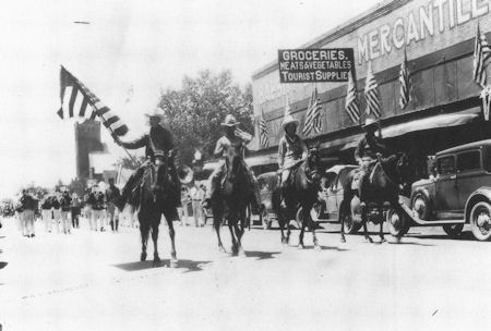 Black and white photo of people on horseback leading a parade down a street lined with American flags and storefronts, including a grocery and mercantile store.