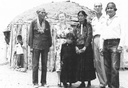 Six adults and one child stand in front of a traditional round wooden structure; another child and adult are partially visible at the entrance.