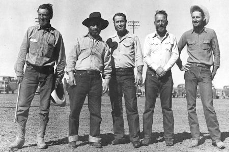 Five men in casual shirts, jeans, and boots stand side by side outdoors on a dirt surface with vehicles in the background.
