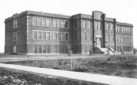 Black and white photo of a large, three-story brick school building with many windows and a central entrance featuring stairs.