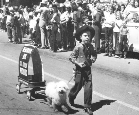 A boy in cowboy attire leads a fluffy dog pulling a small cart with a sign, while a crowd watches from the sidewalk.