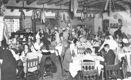 Black and white photo of a crowded restaurant or banquet hall with people dining at tables and decorations hanging from the ceiling.