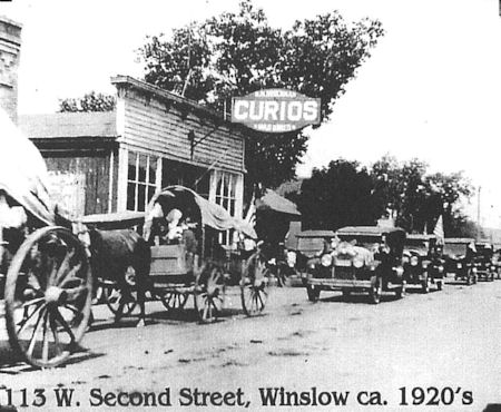 Black and white photo of horse-drawn wagons lined up in front of a store with a "Curios" sign at 113 W. Second Street, Winslow, circa 1920s.