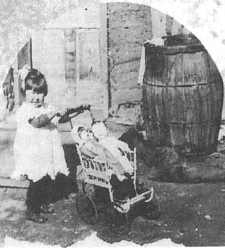 A young girl in a white dress pushes a doll in a small stroller outside near a wooden barrel and a building.