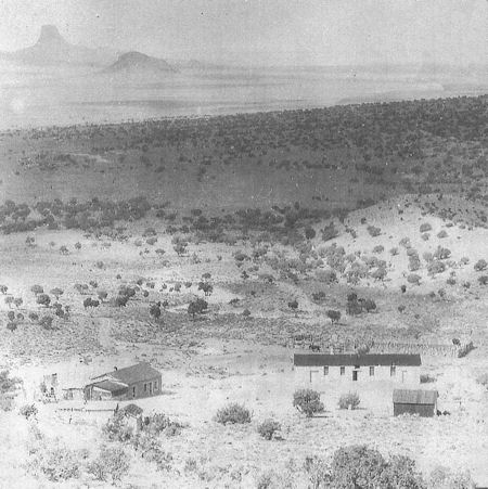 Black and white photo of a rural landscape with scattered shrubs, three small buildings in the foreground, and distant mountains in the background.