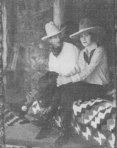 Two people wearing cowboy hats and scarves sit side by side on a patterned blanket, possibly indoors, facing the camera in a black-and-white photograph.