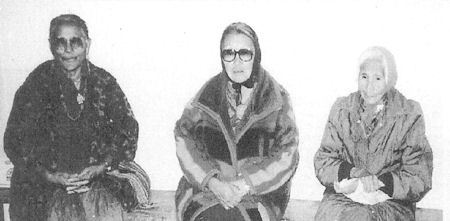 Three elderly women sit side by side on a bench, each wearing glasses and traditional clothing, facing the camera against a plain background.