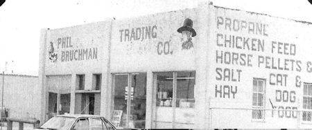Black and white photo of a storefront labeled "PHIL BRUCHMAN TRADING CO." advertising propane, chicken feed, horse pellets, salt, hay, cat and dog food. A car is parked in front.