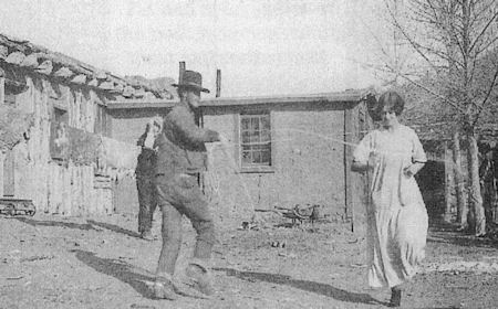 A man and a woman dance outdoors near a rustic building, while another person stands in the background.