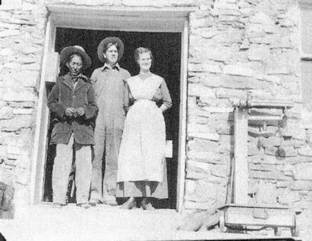Three people stand in the doorway of a stone building. The individuals wear early 20th-century attire, with one woman in a dress and two men in hats and work clothes.