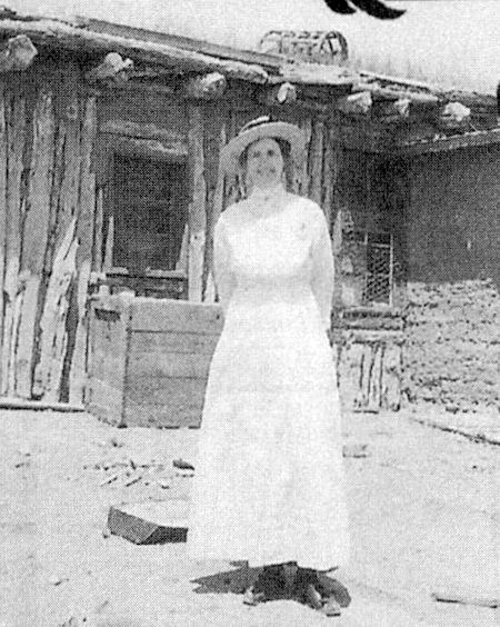 A woman in a long white dress and hat stands in front of a rustic wooden and adobe building, with scattered debris on the ground.