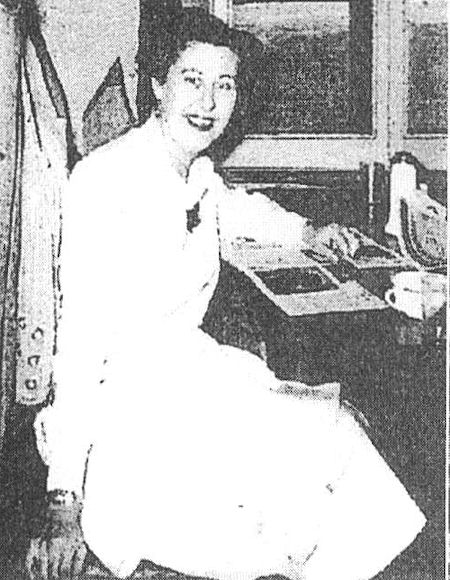 A woman in a light-colored dress sits at a desk, smiling at the camera, with papers and a cup on the desk beside her.