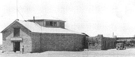 Black and white photo of a large stone building with an attached lower structure and a vintage car parked nearby; a person stands near the entrance.