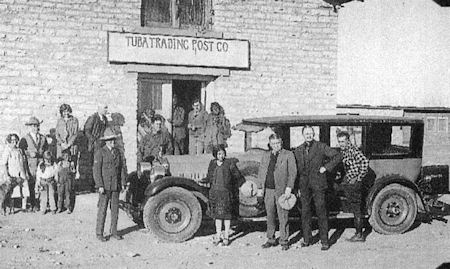 A group of people stands in front of an old car and the "Tuba Trading Post Co" building, posing for a photo in daylight.